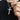 Hand holding a silver-tone cross pendant with engraved text on its side, on a link chain over a black t-shirt